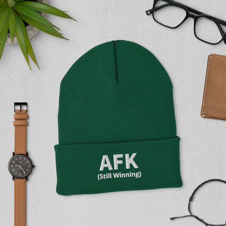 AFK cuffed beanie in green with embroidered text AFK (Still Winning), surrounded by accessories on a table.